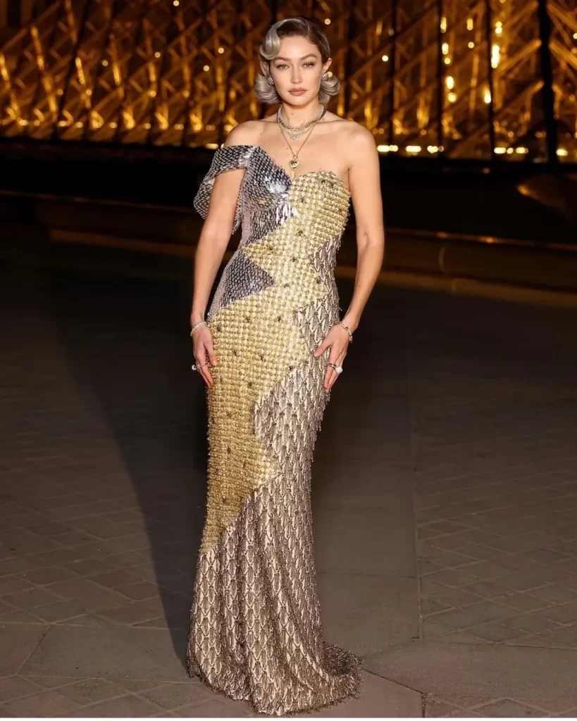 At yesterday's Le Grand Dîner du Louvre in Paris, Gigi Hadid made a striking appearance in a custom Moschino gown. The dress featured a unique design, combining three distinct metallic elements: a silver chainmail segment with an off-the-shoulder sleeve, a middle section adorned with yellow and white rhinestones, and a bronze beaded fringe portion.